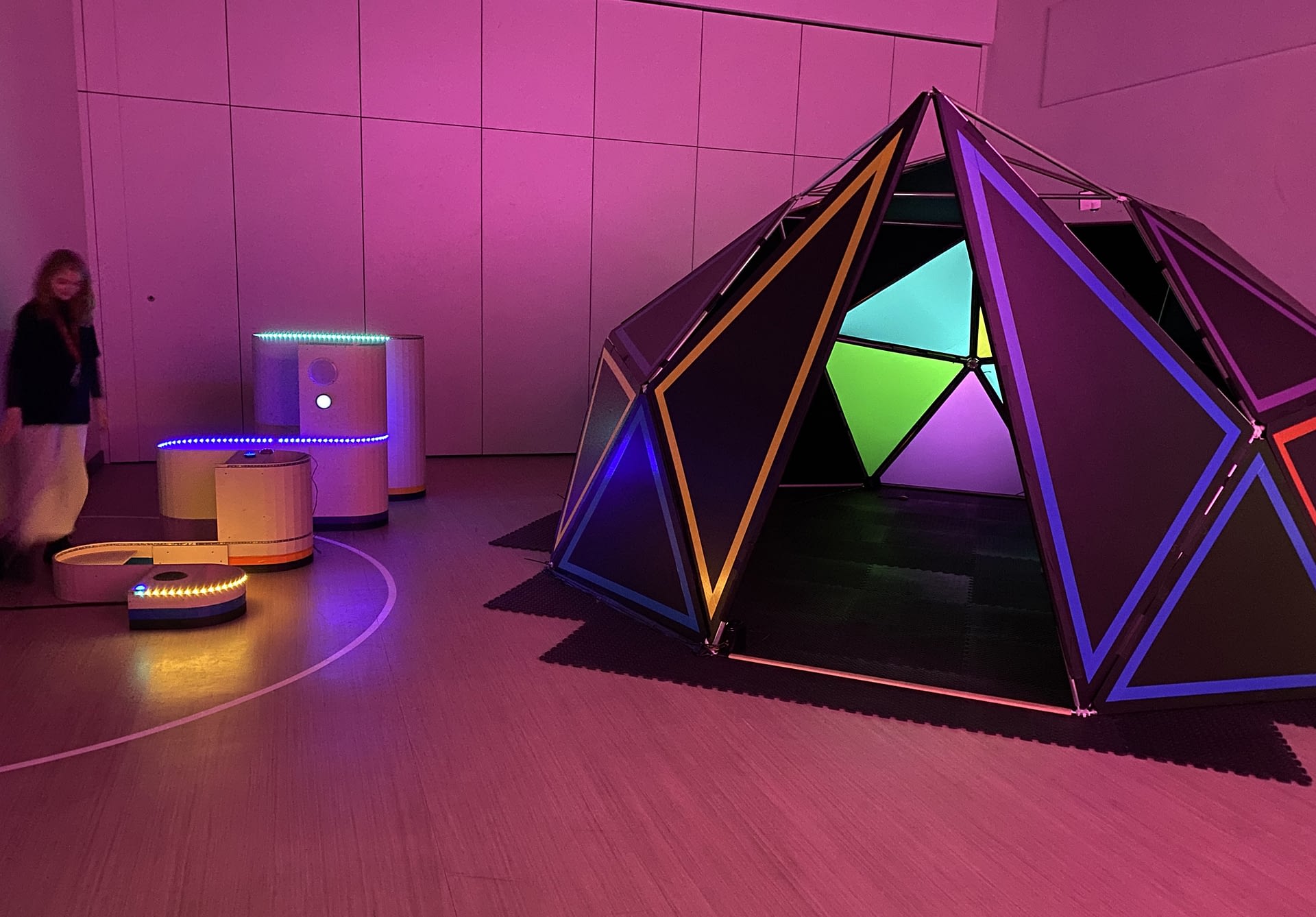 A black geometric dome with colourful panels inside next to a structure with LED lights in the form of a sign wave in a room with pink lighting